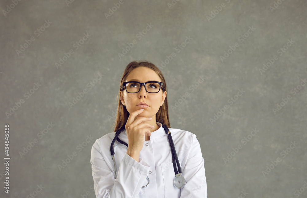 Portrait of a dreamy young female doctor with a pensive expression on a gray background. Pensive doctor or nurse with glasses, medical gown and stethoscope looking up touching his chin. Banner. Portrait of a dreamy young female doctor with a pensive expression on a gray background. Pensive doctor or nurse with glasses, medical gown and stethoscope looking up touching his chin. Banner.