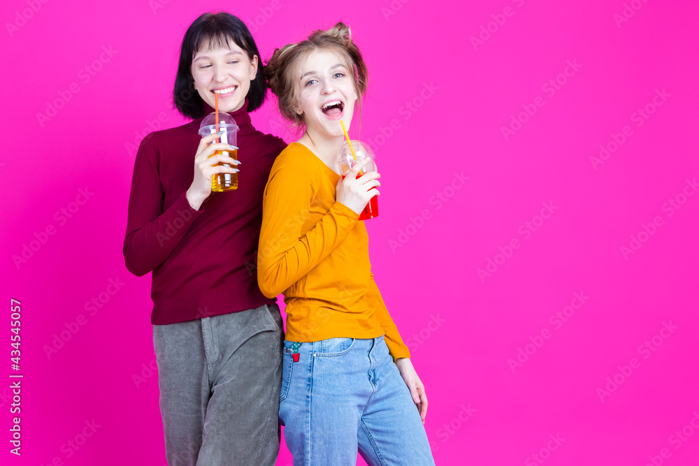 Two Laughing Smiling Caucasian Girls Together with Cups Filled With Colorful Juice or Smoothie Posing Over Pink Background. Two Laughing Smiling Caucasian Girls Together with Cups Filled With Colorful Juice or Smoothie Posing Over Pink Background.