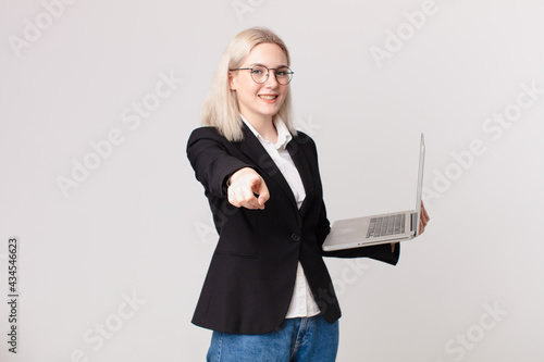 blond pretty woman pointing at camera choosing you and holding a laptop