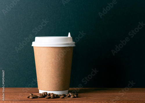 Disposable paper cup with hot coffee and roasted coffee grains on wooden table in front of black wall. Space for text