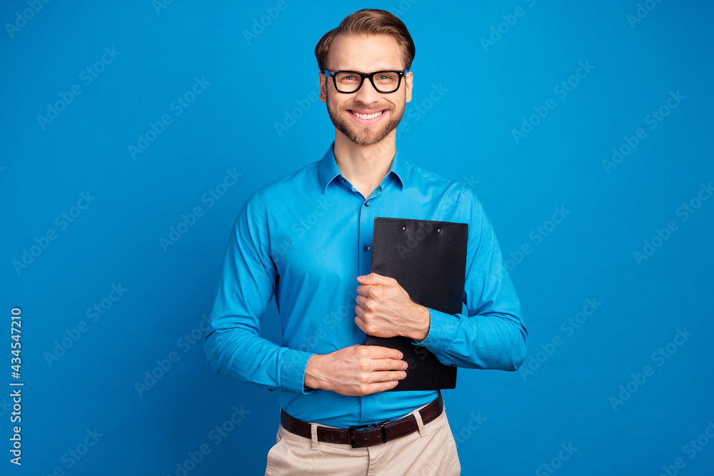 Photo of young business man happy positive smile hold clipboard organizer isolated over blue color background Photo of young business man happy positive smile hold clipboard organizer isolated over blue color background