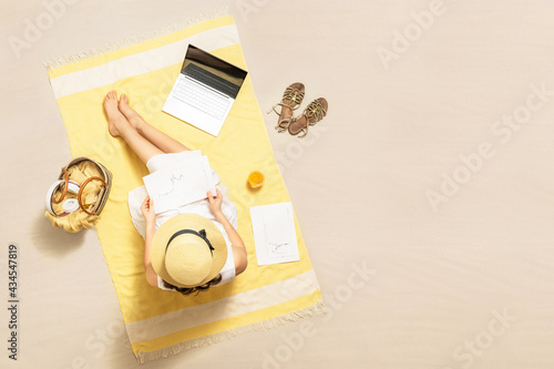 Businesswoman freelancer drinking juice sitting  on beach towel of sand beach and working on laptop with graphics and charts. Summer vacation. Aerial view