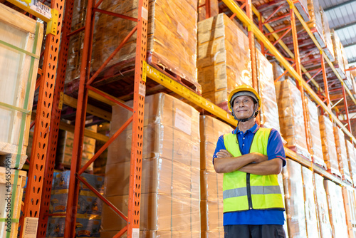 Working at warehouse.Portrait worker asian man in safety with yellow hardhat standing with crossed arms and smilling.Male asia engineer at large Warehouse works with goods