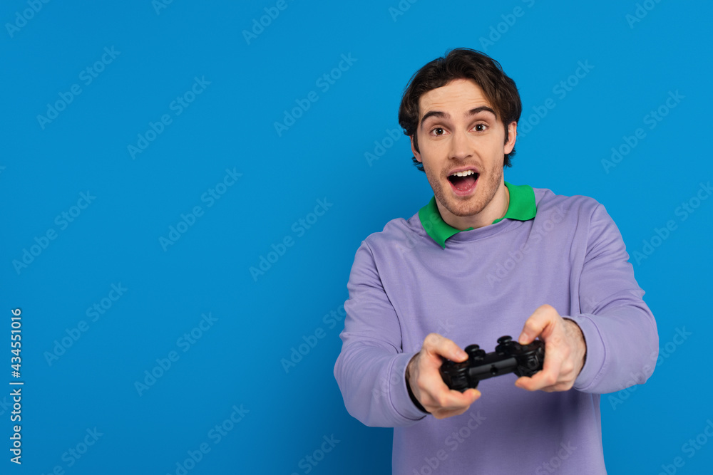 KYIV, UKRAINE - MARCH 22, 2021: young man holding joystick and looking at camera isolated on blue KYIV, UKRAINE - MARCH 22, 2021: young man holding joystick and looking at camera isolated on blue