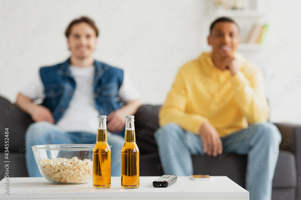 bottles of beer, TV remote and popcorn on blurred background with two interracial friends sitting on couch bottles of beer, TV remote and popcorn on blurred background with two interracial friends sitting on couch