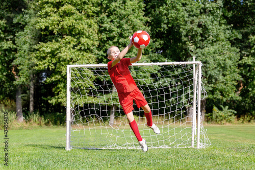 A young soccer player-goalkeeper in a red uniform catches the ball, defending the goal. Children's sports, physical activity concept