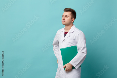 Young handsome doctor in a white coat holding a green folder isolated on a blue background.