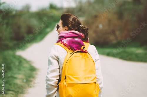 Back view of woman traveller with yellow backpack at the beginning of a country road in bright vintage colors. Concept: adventure, travel post pandemic, countryside roads, empty roads