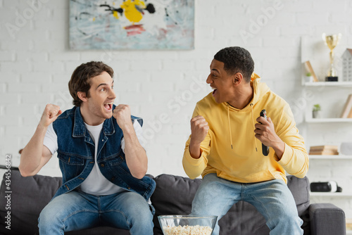 excited interracial friends watch tv and eating popcorn in modern loft