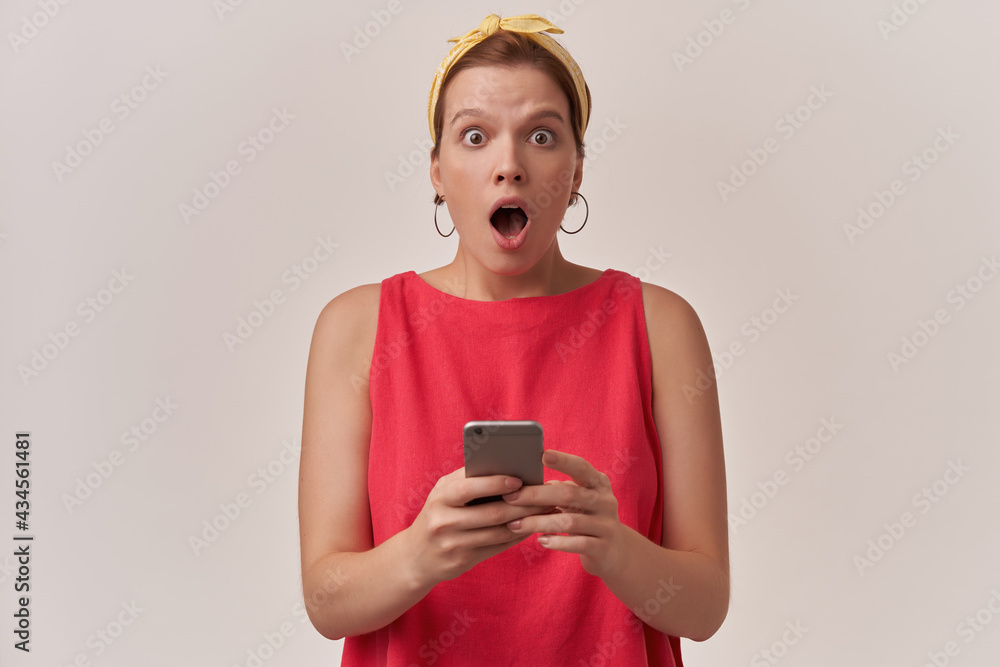 Image of brown eyed charming woman 20s with natural makeup and earrings wearing stylish trendy red dress and yellow bandana posing against white background surprised looking at you hold phone in arms Image of brown eyed charming woman 20s with natural makeup and earrings wearing stylish trendy red dress and yellow bandana posing against white background surprised looking at you hold phone in arms