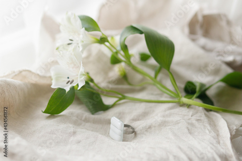 Stylish modern white square ring and alstroemeria beautiful flower on linen textile, copy space. Unusual fashionable fused glass ring. Contemporary gift