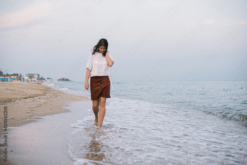 Carefree hipster woman walking barefoot on sandy beach with sea waves in the evening, enjoying tranquil moment. Casual young female  relaxing on seashore at resort. Mindfulness