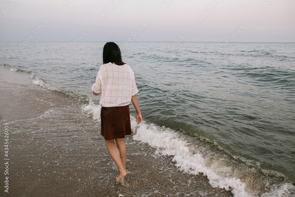 Carefree hipster woman walking barefoot in cold sea waves on sandy beach, enjoying tranquil evening. Casual young female relaxing on seashore at resort, back view. Calm and Mindfulness