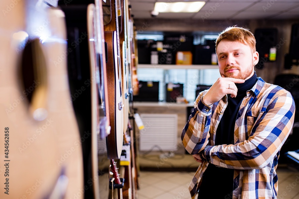 redhaired bearded handsome man in brown plaid casual shirt choosing a guitar in a music store