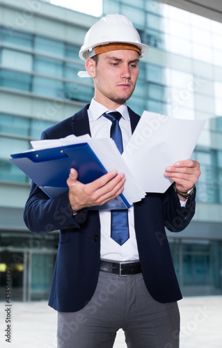 Smiling builder in suit and hat with folder is exploring documents with project near the building. photo