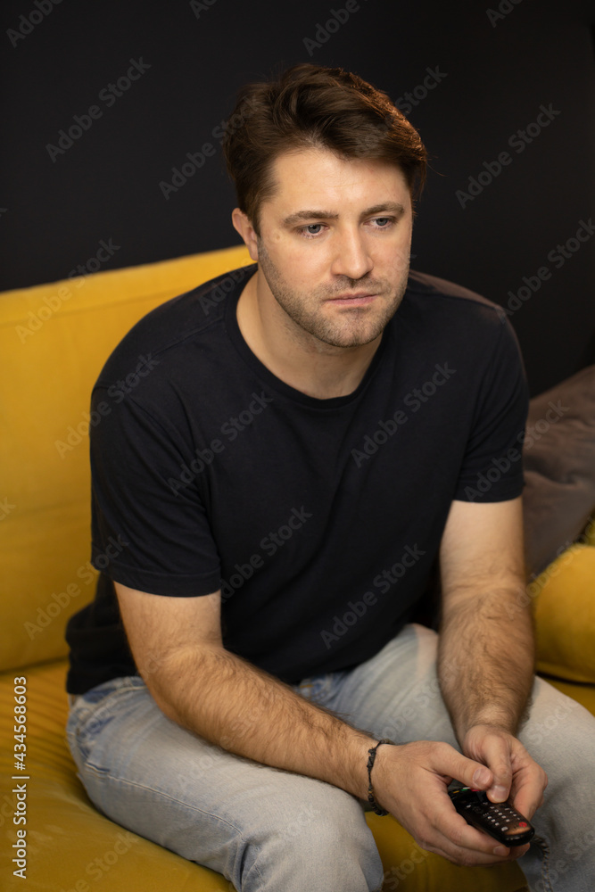 Lockdown Pastime boredom concept. Young unshaven handsome guy sitting on comfy yellow sofa clicks remote control watching movie on TV or youtube videos. Boring Evening entertainment at Home Alone, Lockdown Pastime boredom concept. Young unshaven handsome guy sitting on comfy yellow sofa clicks remote control watching movie on TV or youtube videos. Boring Evening entertainment at Home Alone,