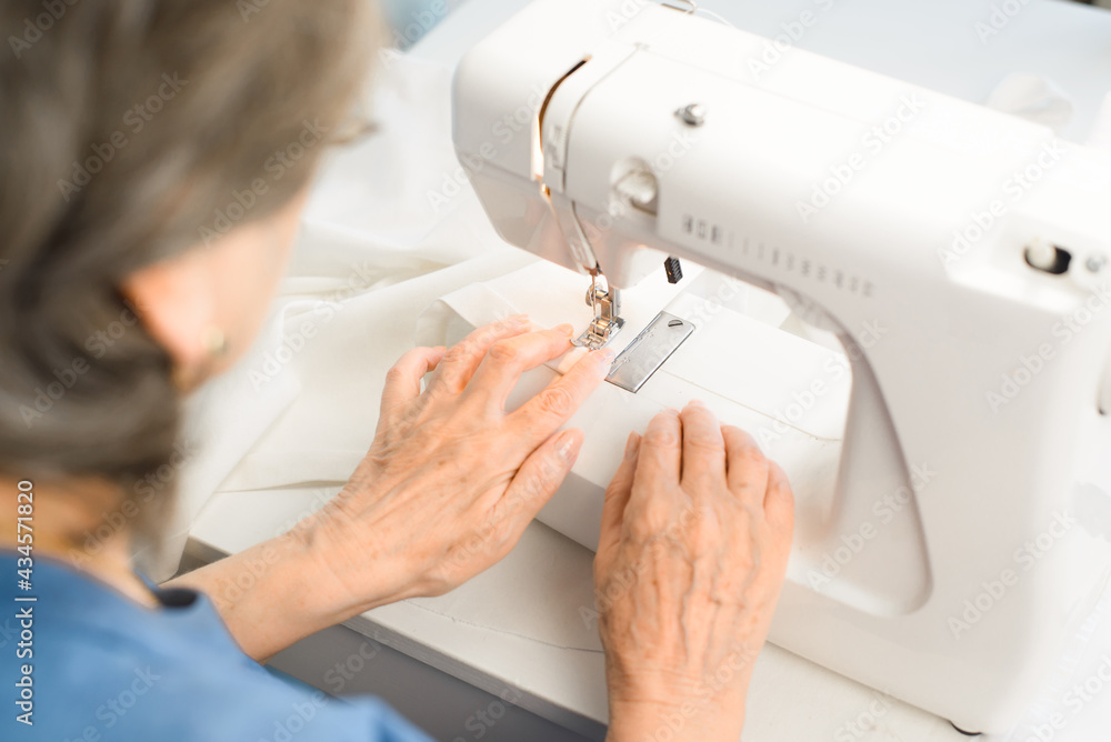 Woman aged using a sewing machine at home. Senior woman dressmaker sews something from white fabric, view from the back. Hobby or work at home for a pensioner. Selective focus on hands