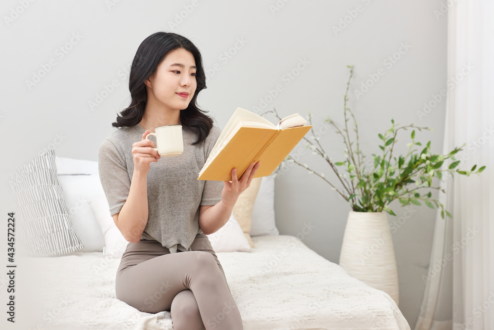 침실을 배경으로 침대에 앉아서 커피 또는 차를 마시며 책을 보고있는 젊은 아시아 한국 여자 모델