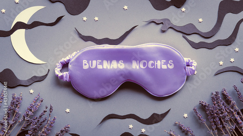 Sleep mask with dry lavender flowers. Text Buenas Noches means Good night in Spanish. Silver grey paper background with black clouds and Moon. Aromatherapy, scented herbs. Creative top view, flat lay.