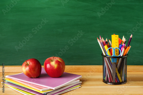Back to school background. Pens, pencils, rulers, scissors in a glass, two red apples, a stack of notebooks on the background of a green school board, closeup, copy space.