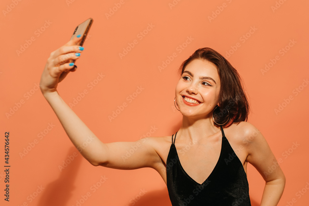positive young woman taking selfie and smiling on orange background positive young woman taking selfie and smiling on orange background