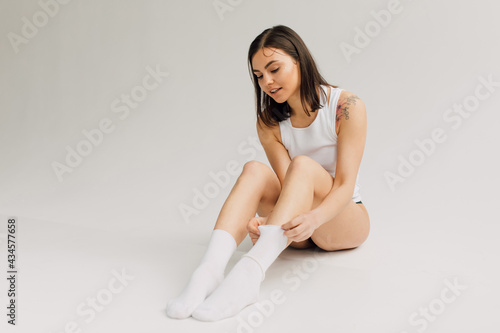 young woman in white top and black panties adjusting socks on grey background