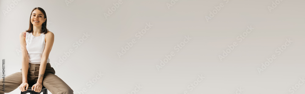 young smiling woman in white top and beige trousers sitting on stool on grey background, banner young smiling woman in white top and beige trousers sitting on stool on grey background, banner