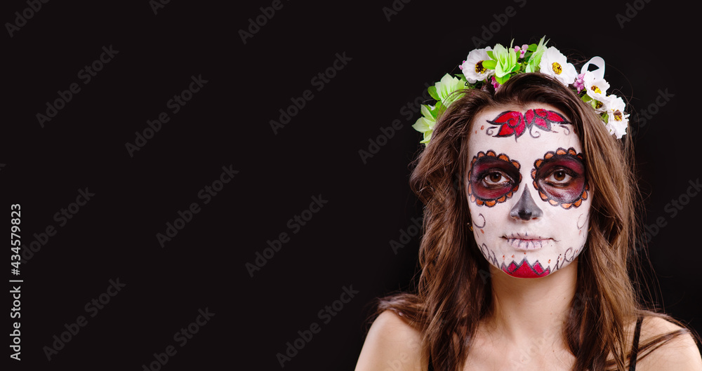 A young girl with dark hair and sugar skull makeup with flowers on her head looks into the camera on a black background. Panoramic stretched image for banner