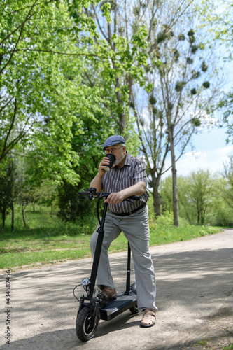 A positive, charismatic elderly man on an eco scooter went for a walk in the park. Drinks a drink from an eco paper cup. Healthy lifestyle of the elderly.
