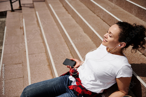 Young beautiful woman leaning on the steps holds a smartphone in her hands and listens to music on headphones