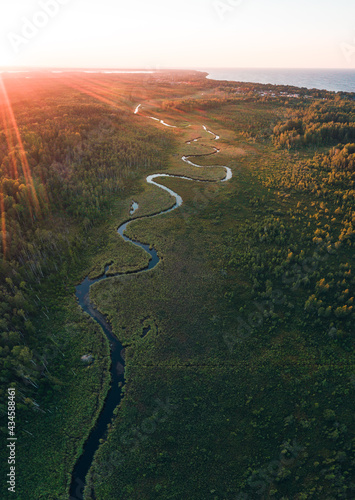 Fototapeta Naklejka Na Ścianę i Meble -  Winding river flowing through forest towards sea. Aerial sunset. Sunlight rays. Baltic sea, Latvia.