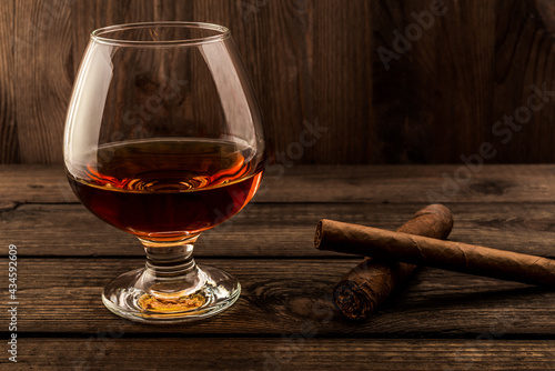 Glass of brandy and two different cuban cigars on a old wooden table. Focus on the cuban cigars