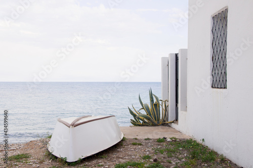 Upside down white boat on the beach, next to a white house and a green plant. Calm sea in the background. Altea, Spain