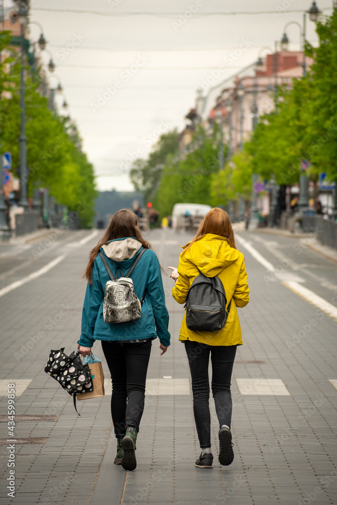 Couple of young friends girls walking and chatting in the middle of the main street in the city with backpacks and umbrella before or after the rain