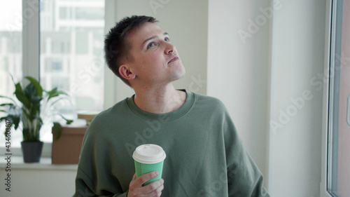A man drinks coffee from a cardboard cup near the window