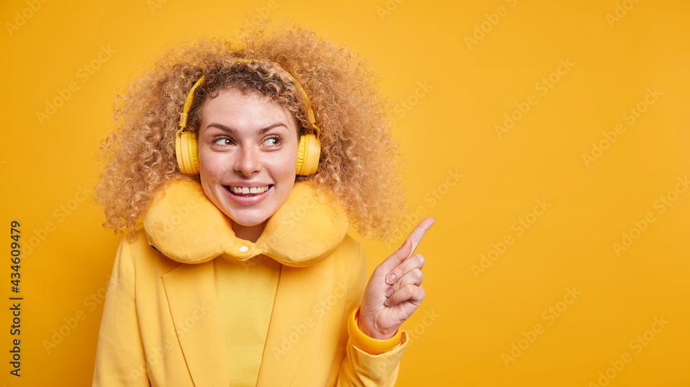 Good looking positive woman with bushy curly hair attracts your attention to copy space demonstrates product points away uses wireless headphone enjoys playlist wears neck pillow for comfort Good looking positive woman with bushy curly hair attracts your attention to copy space demonstrates product points away uses wireless headphone enjoys playlist wears neck pillow for comfort