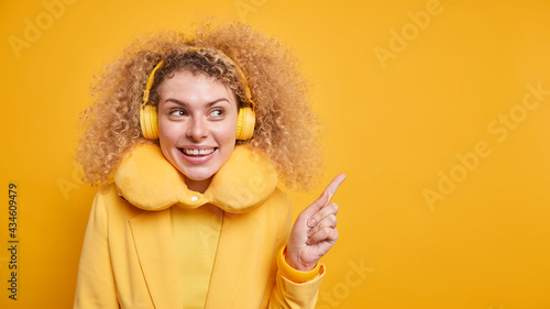 Good looking positive woman with bushy curly hair attracts your attention to copy space demonstrates product points away uses wireless headphone enjoys playlist wears neck pillow for comfort © wayhome.studio 