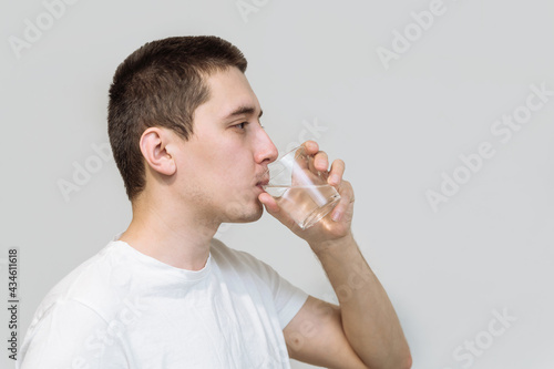 a young man in a white T-shirt drinks pure water in a clear glass glass on a white background. health concept