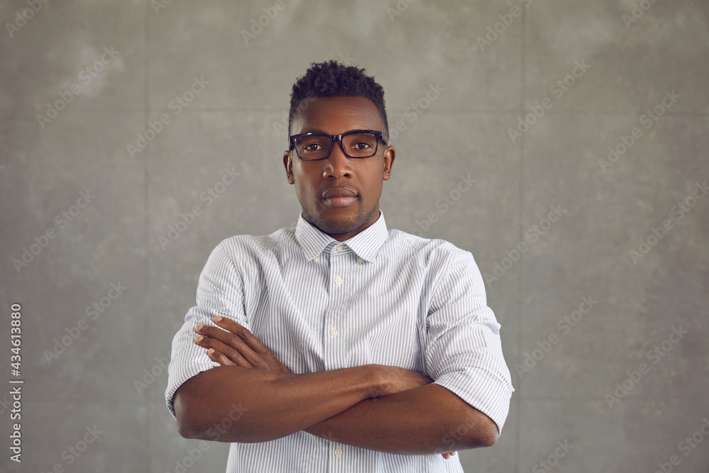 Studio portrait of serious handsome young black man in white shirt and glasses standing arms crossed looking at camera. Profile picture of African American salesperson isolated on gray background
