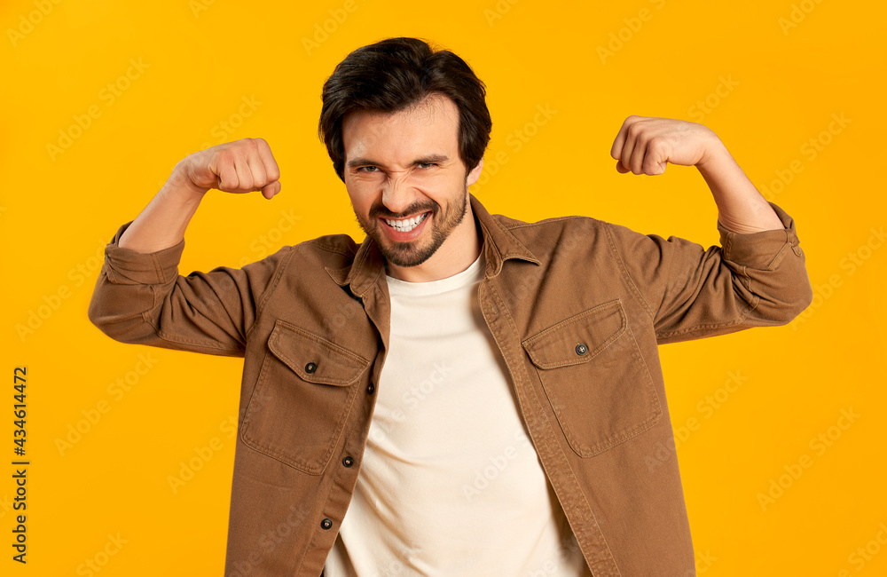 Young bearded man shows a gesture of strength, muscles on his arms isolated on orange background. Young bearded man shows a gesture of strength, muscles on his arms isolated on orange background.