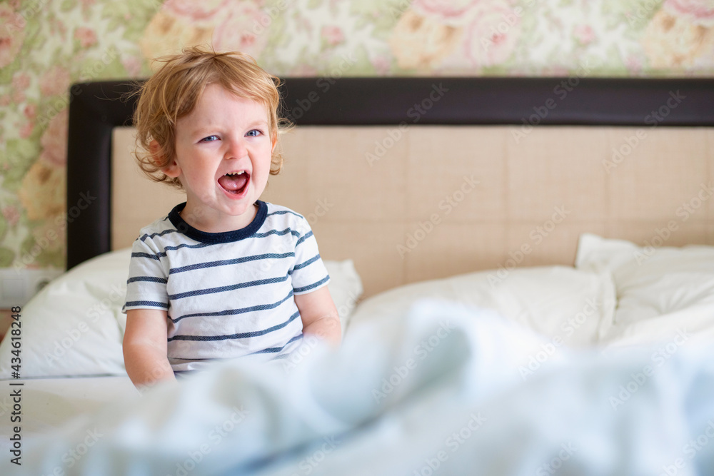 Cute laughing child boy sits in bed. Boy plays at home. Child in a sunny bedroom Cute laughing child boy sits in bed. Boy plays at home. Child in a sunny bedroom