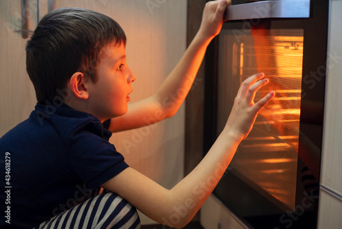 A child is sitting near the oven in the kitchen and waiting. Curious boy is watching through the glass of kitchen oven. Baking pizza, muffins , cupcakes or cookies