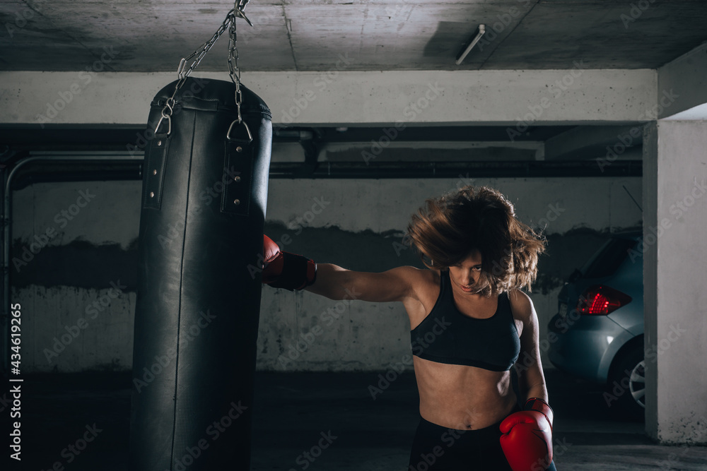 Profile view of a strong young woman punching a boxing bag inside the garage