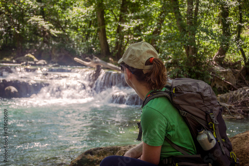 Rear view of a blonde Caucasian woman with a hat and a backpack relaxing and enjoying the scenery at a waterfall in the forest. Sentierelsa  Colle di Val d Elsa - Siena  Tuscany  Italy