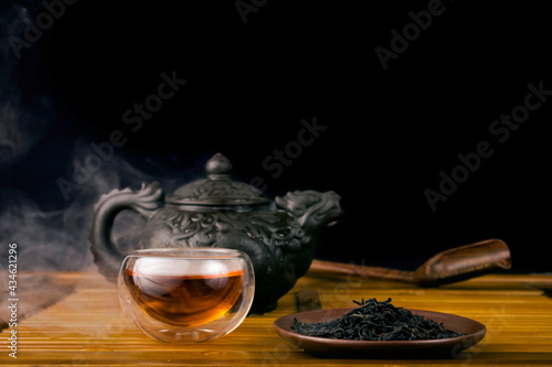 Chinese tea ceremony. Clay teapot with red tea Lapsang souchong on a black background, heap of dry tea leaves and glass thermo cup of hot tea with vapour. photo