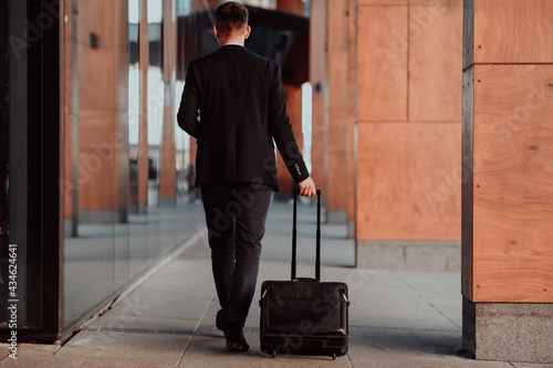 Going to airport terminal. Confident businessman traveler walking on city streets and pulling his suitcase drinking coffee and speaking on smartphone