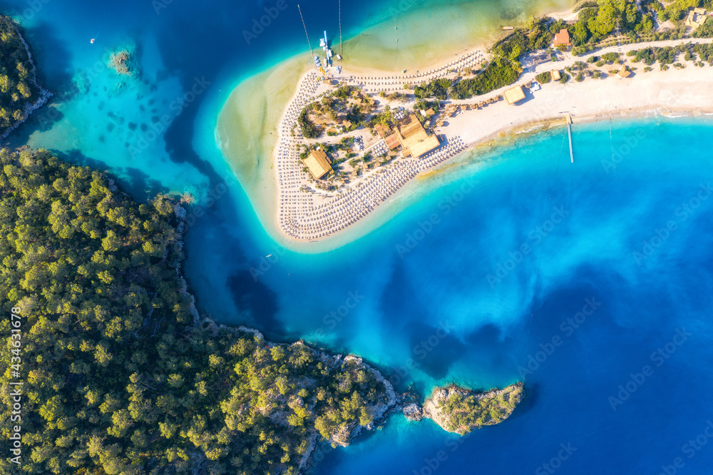 Aerial view of sandy beach with umbrellas, green trees, mountain at sunset in summer. Blue lagoon in Oludeniz, Turkey. Tropical landscape with sea bay, island, white sandy bank, blue water. Top view