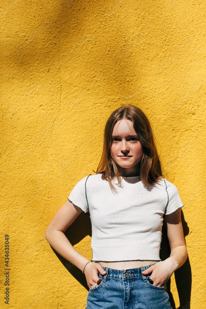 Teenage girl standing next to the yellow concrete wall. Candid portrait in sunny day. City street life authentic people. Young female in urban outfit