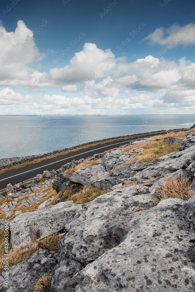 Small asphalt road by the ocean through rough stone terrain, County Clare, Ireland. Blue cloudy sky, Galway bay. Beautiful Irish nature, vertical image