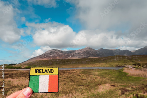 Tourist holding badge with sign Ireland and National flag of Ireland in focus. Mountains out of focus. Connemara, county Galway, Travel and tourism memory concept